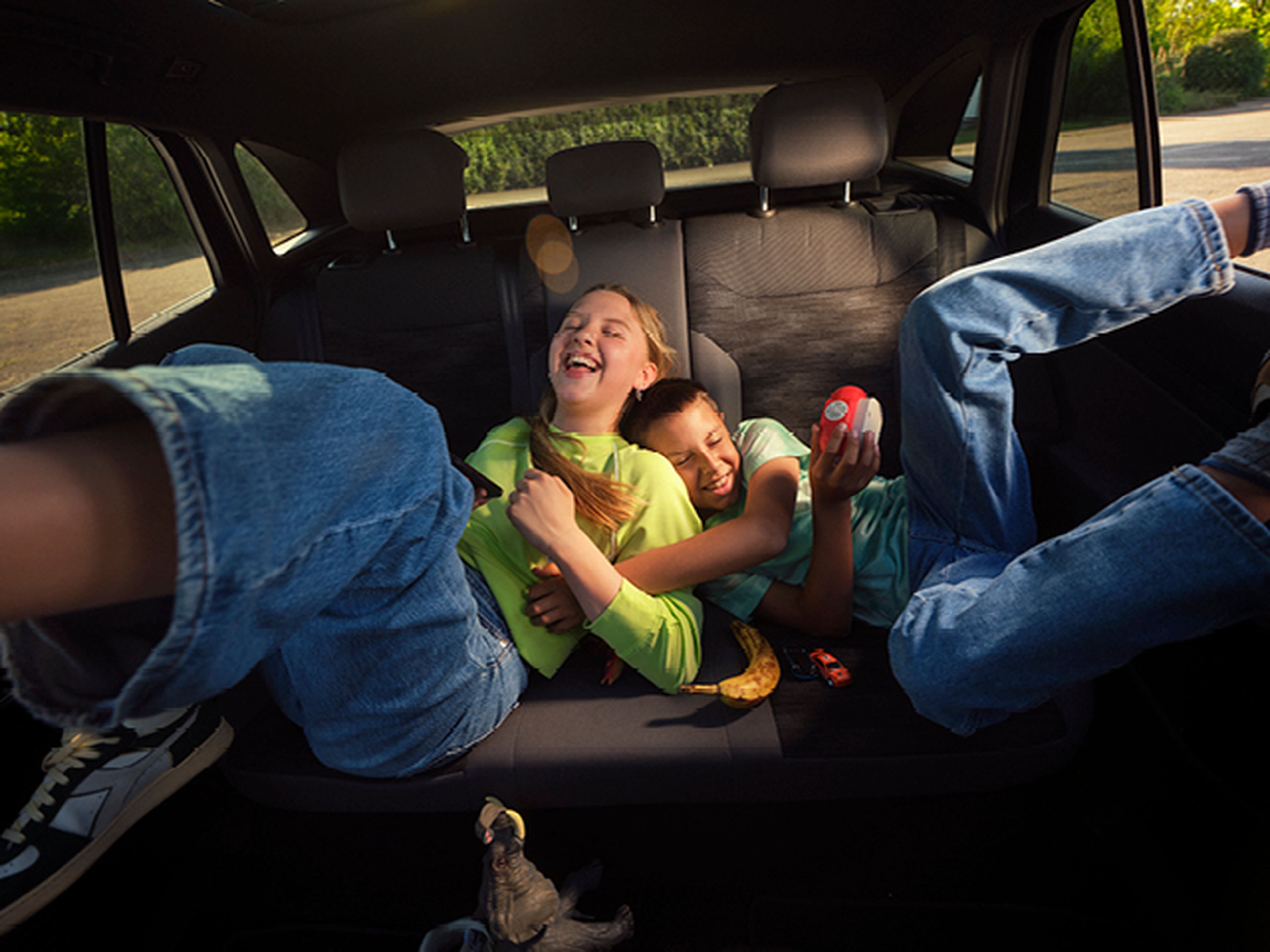 Twee kinderen liggen giechelend op de achterbank van een auto. De jongen knuffelt het lachende meisje in een felgroene trui, terwijl hij een stuk speelgoed vasthoudt.