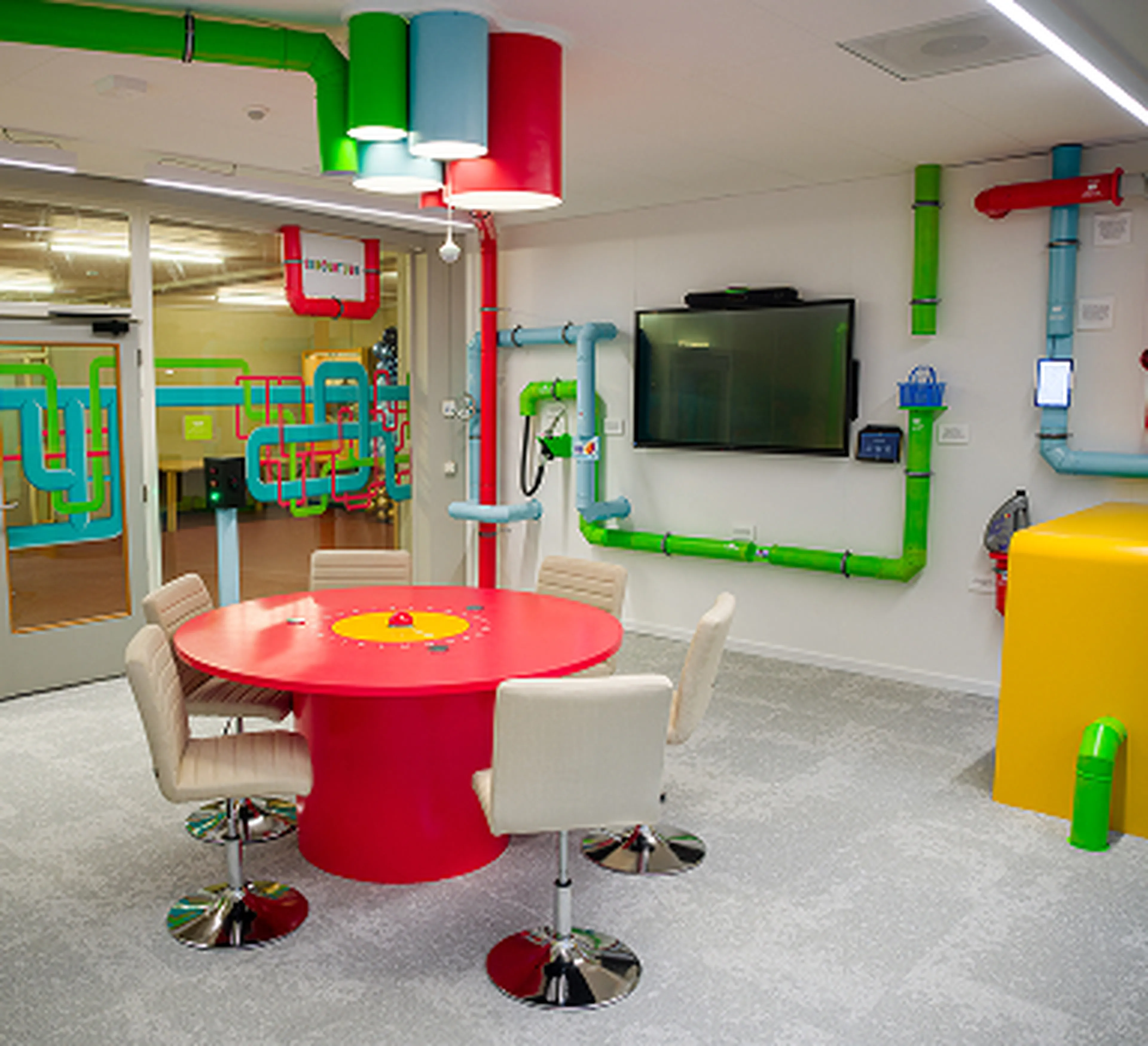 Playfully designed meeting room at the Q8 headquarters in Rotterdam featuring a bright red round table, white chairs, and colorful pipes on the ceiling and walls. A TV screen is mounted centrally on the wall.