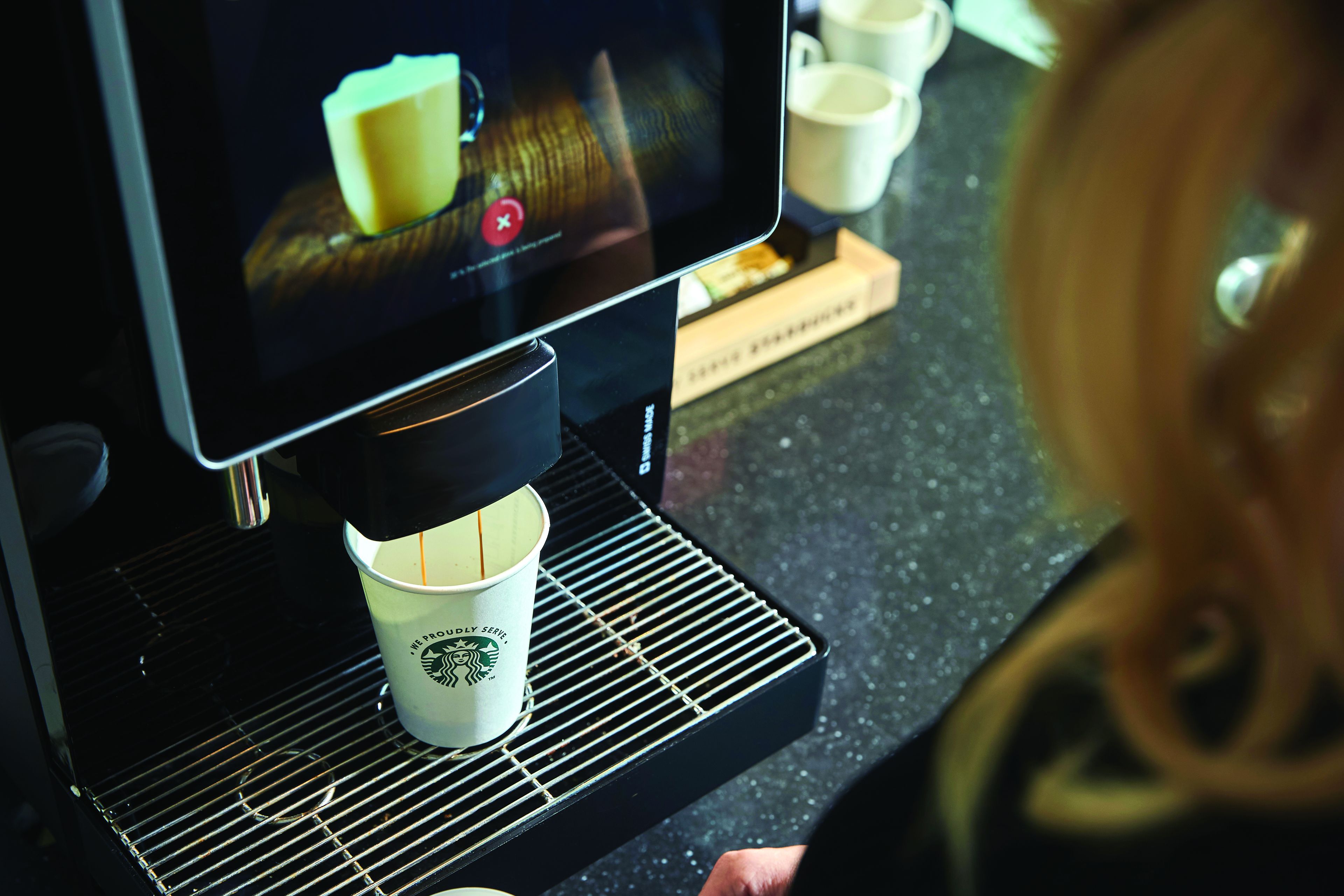 Starbucks-koffiemachine die een warme drank bereidt in een papieren beker, met een touchscreenbediening en een persoon op de achtergrond bij een Q8 tankstation.