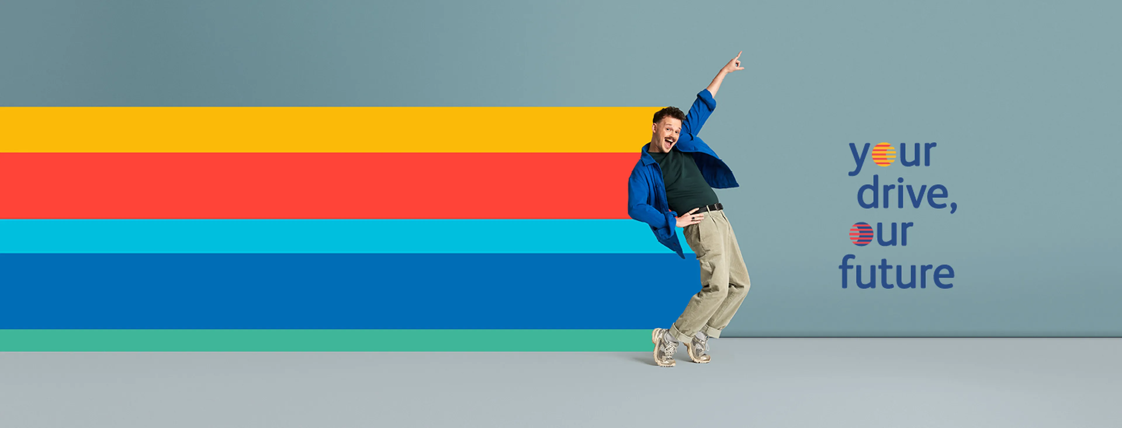 Een persoon in een expressieve pose staat voor een effen blauwe achtergrond en draagt een felblauw overhemd, een donkergroen T-shirt, beige broek en sneakers.