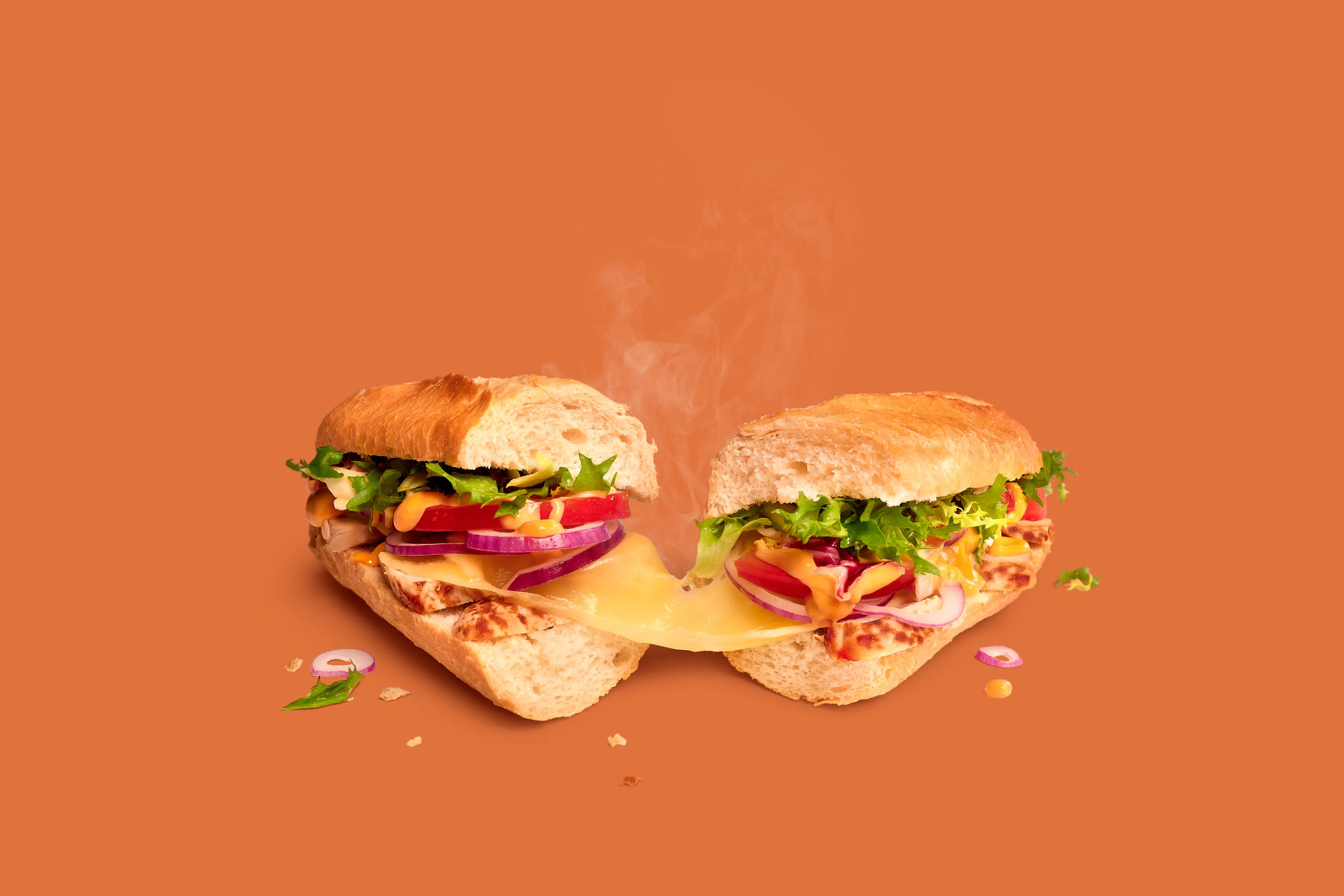 Broodje Chicken Samouraï Remix van Panos, doormidden gesneden, met kip, kaas, groente en saus op oranje achtergrond.