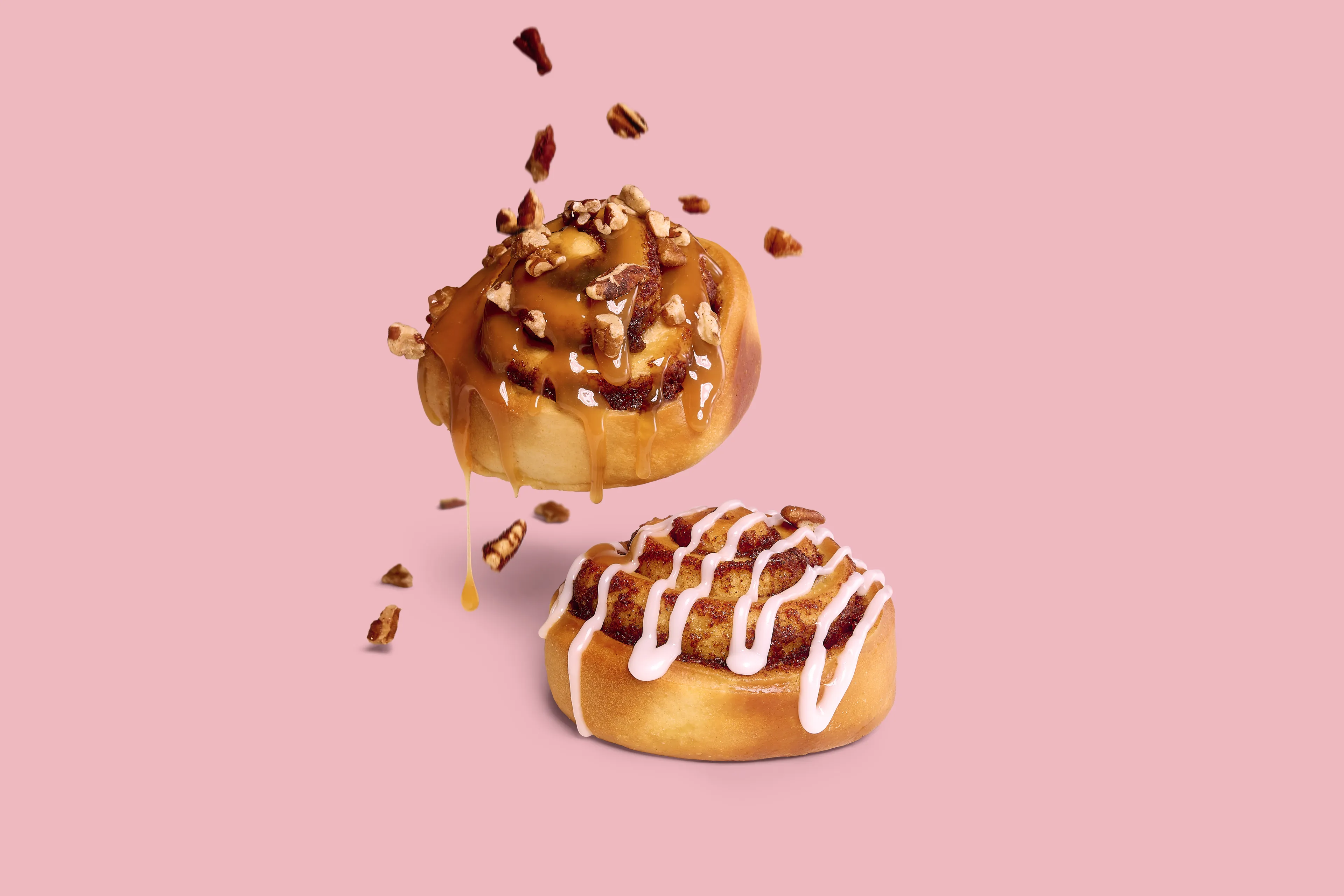 Twee cinnamon rolls van Panos op roze achtergrond, met karamel, pecannoten en wit glazuur.