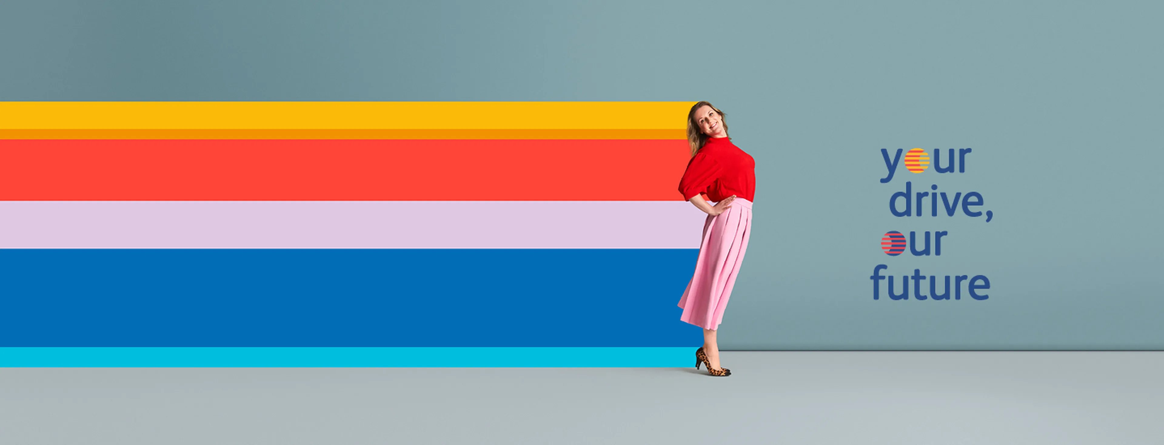A woman is playfully posing with a slight backward lean against a solid blue background. She is wearing a red top, a pink pleated mid-calf skirt, and leopard-print heels.