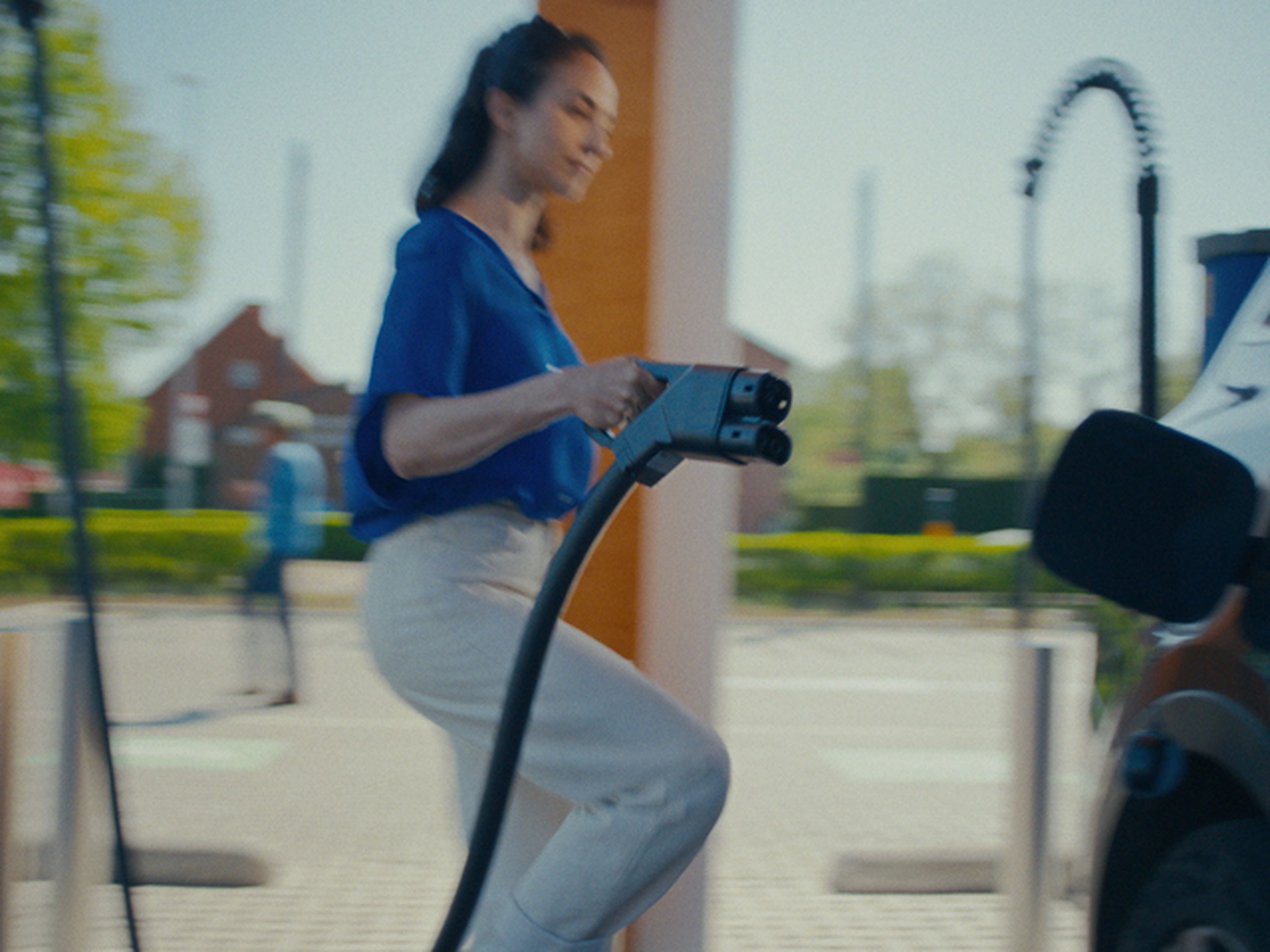 Een vrouw in een blauwe blouse en lichtgekleurde broek loopt naar een elektrische auto terwijl ze een laadkabel vasthoudt. Ze staat op het punt om de auto aan te sluiten bij een Q8 electric laadstation.