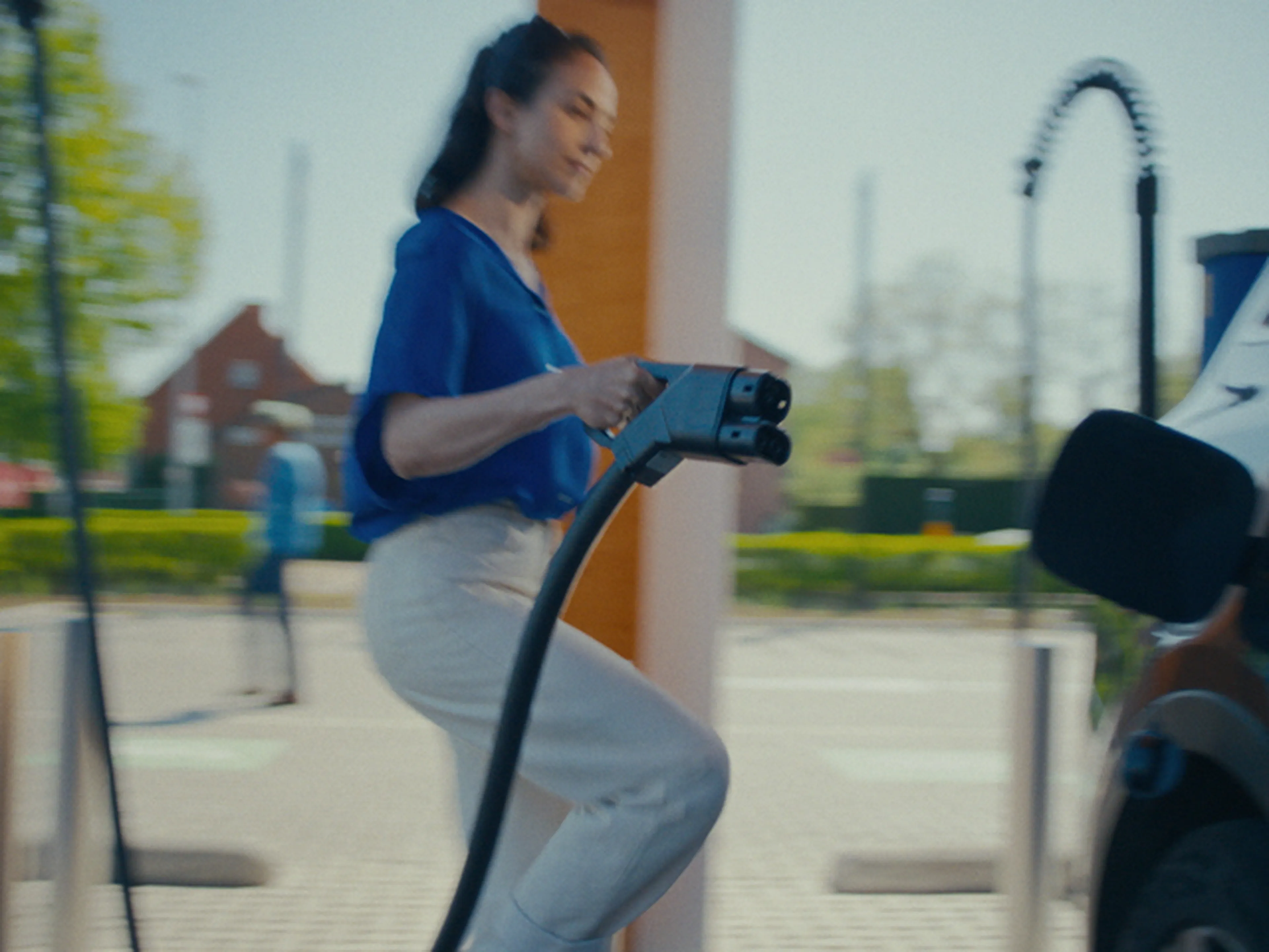 Een vrouw in een blauwe blouse en lichtgekleurde broek loopt naar een elektrische auto terwijl ze een laadkabel vasthoudt. Ze staat op het punt om de auto aan te sluiten bij een Q8 electric laadstation.
