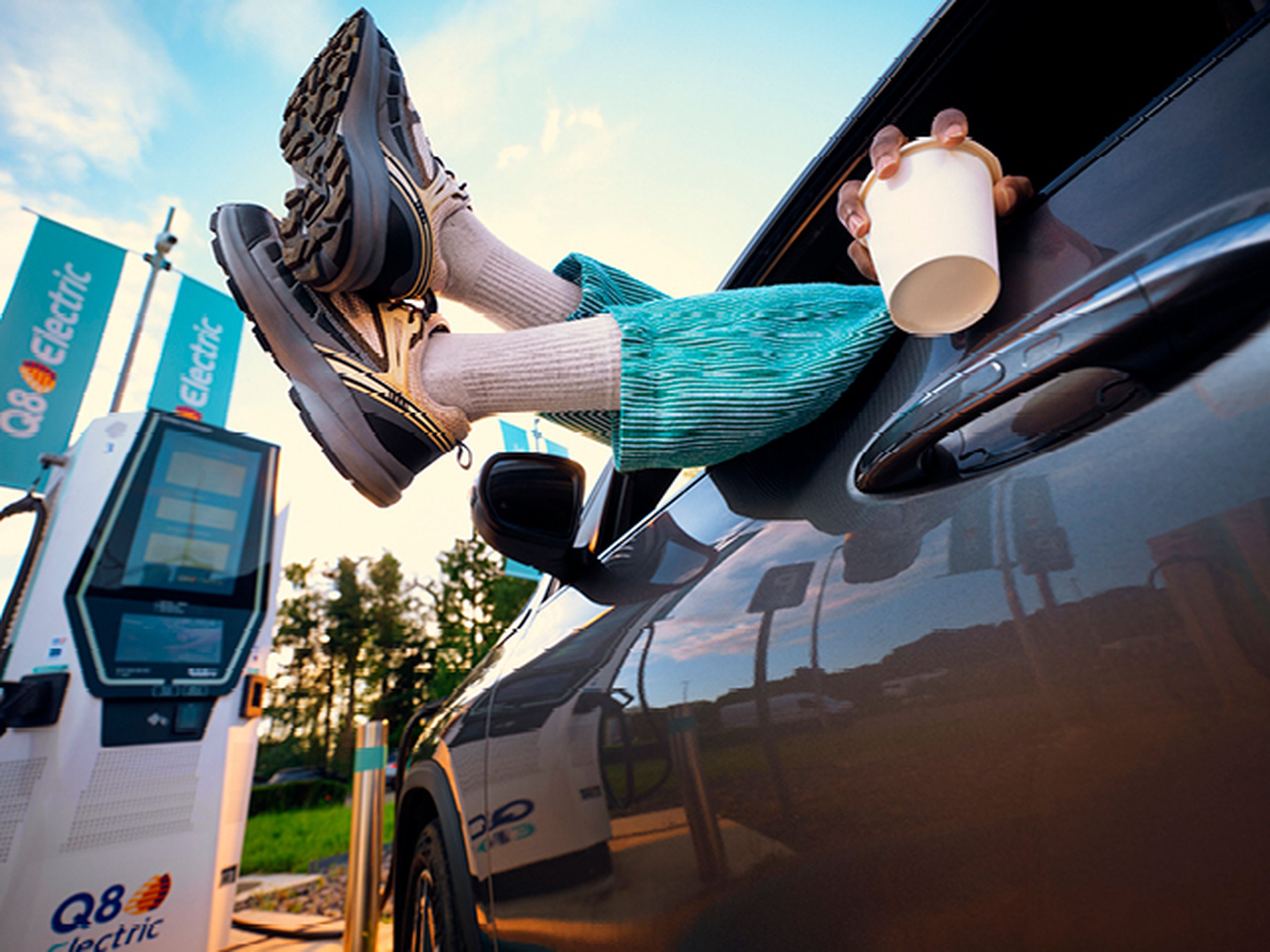 Feet out of car window, person drinking coffee at Q8 Electric charging station.