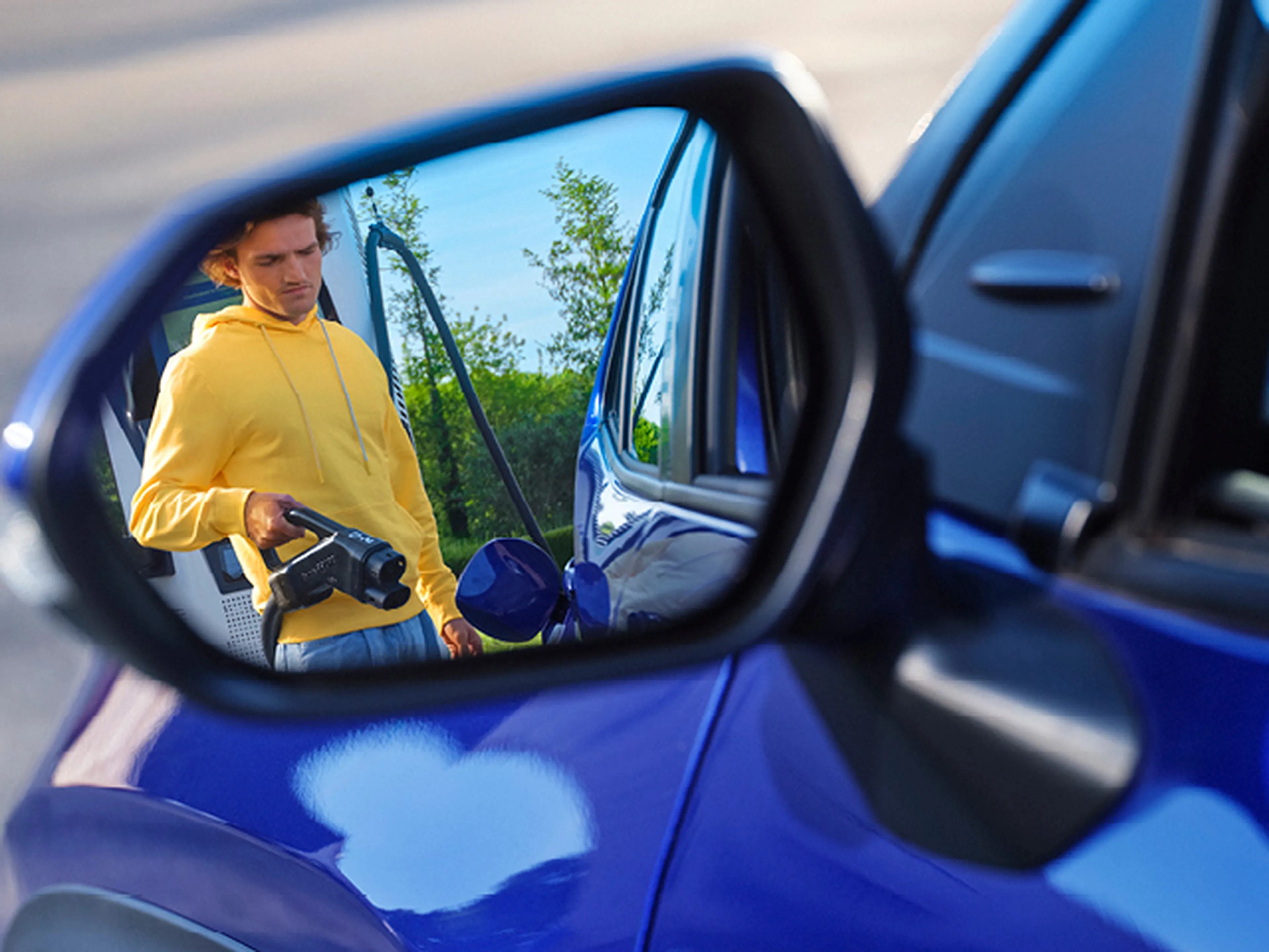 Man met laadkabel reflecteert in de zijspiegel van een blauwe auto bij Q8 electric.