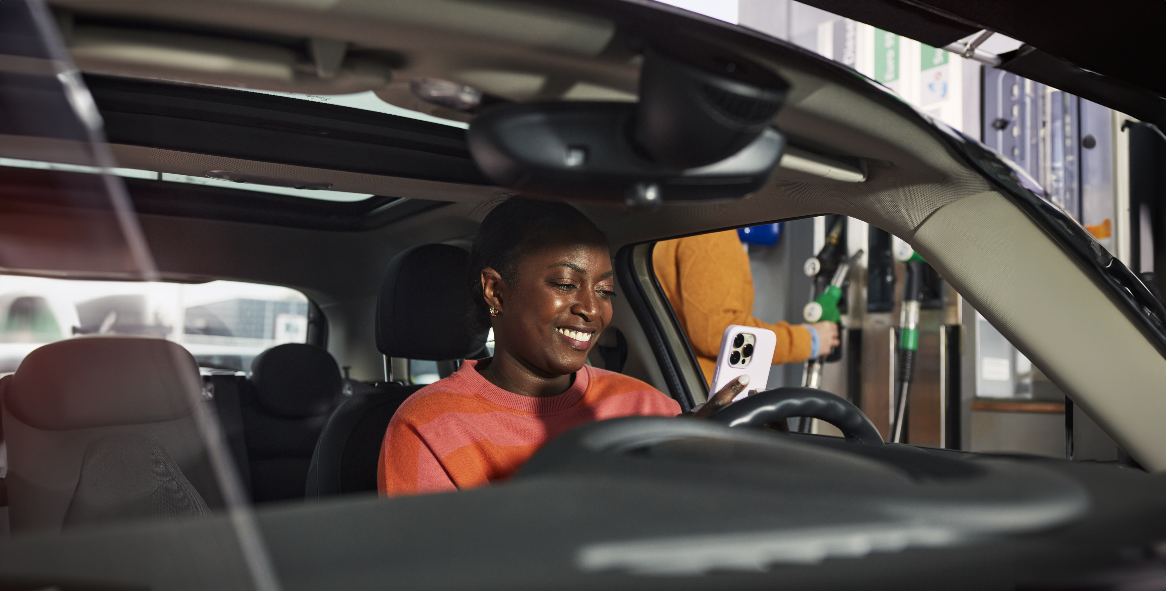 Vrouw met oranje trui kijkt op haar witte smartphone en zit in haar auto.