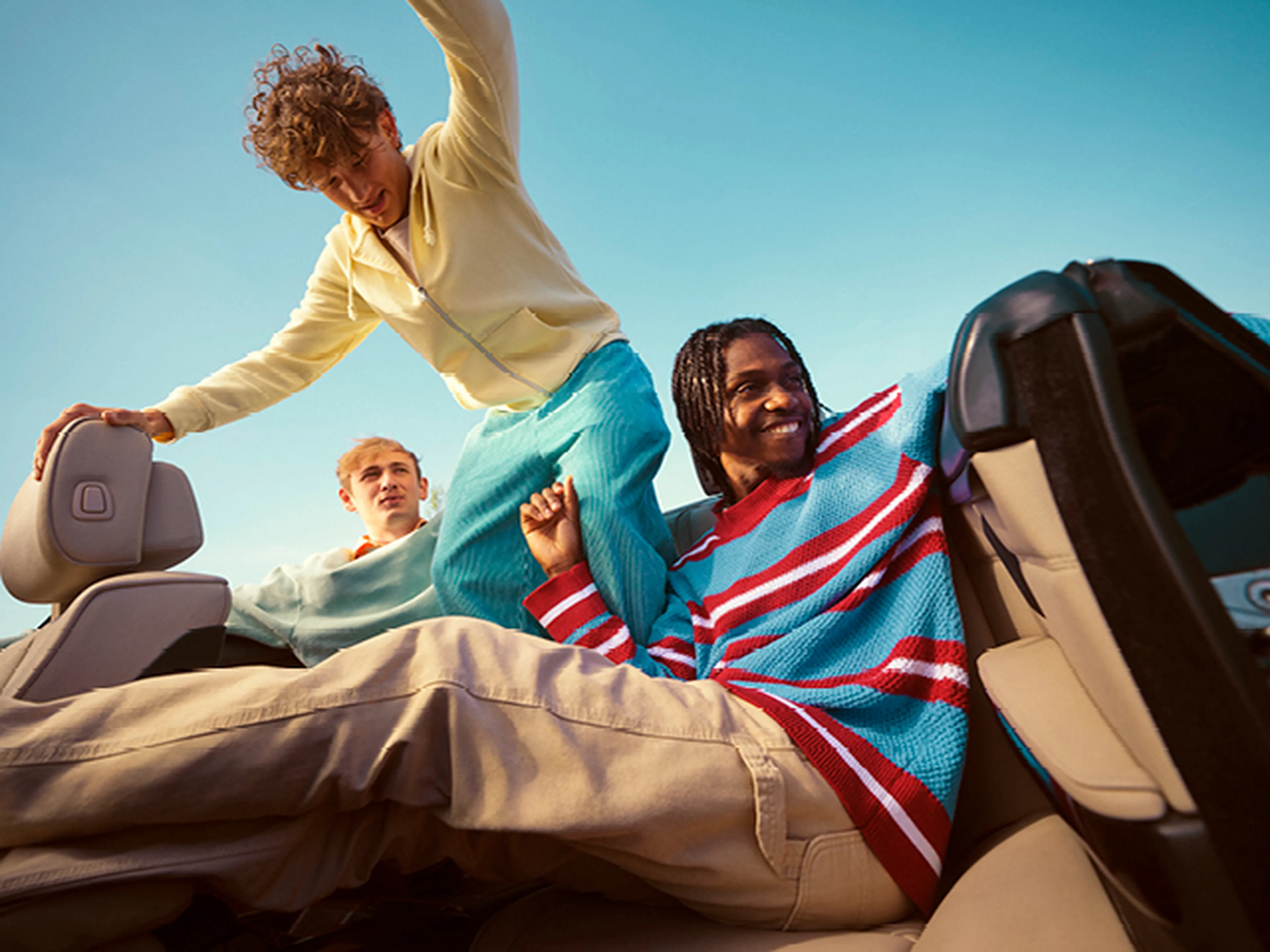 Drie vrienden genieten van een zonnige dag in een cabriolet. Eén persoon klimt over de achterbank, een ander zit ontspannen met een brede glimlach, en de derde kijkt opzij.