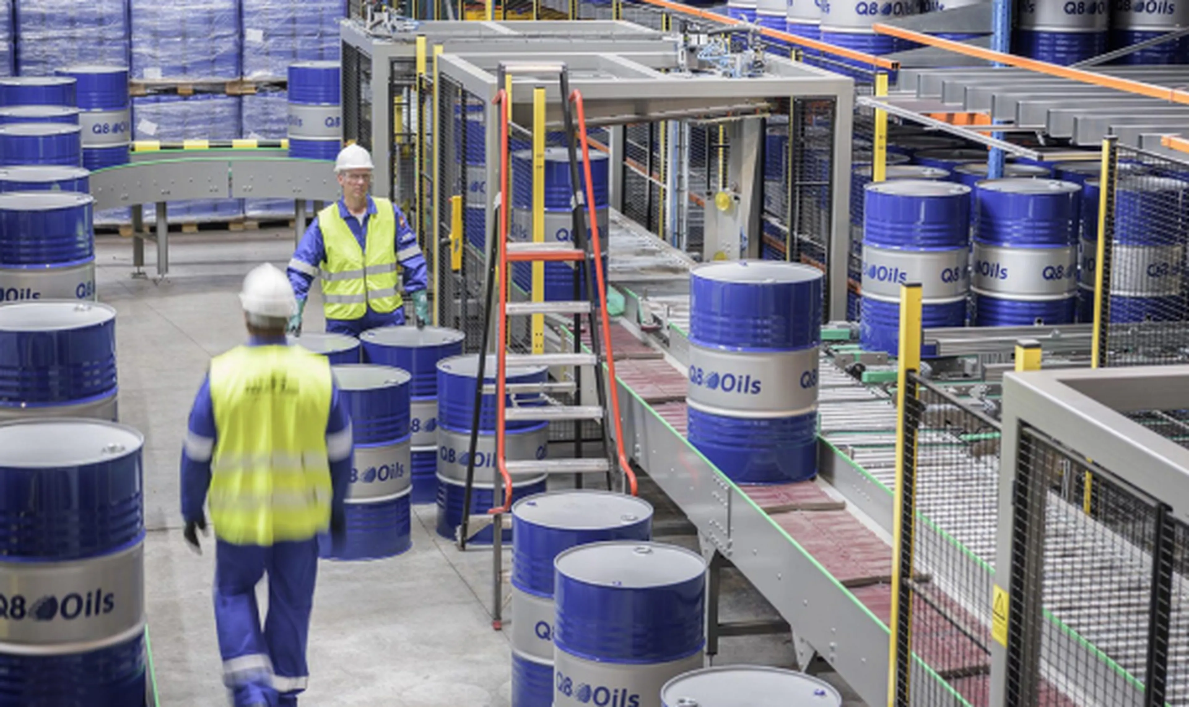 Interior view of the Q8Oils blending plant where blue Q8Oils barrels are positioned on a conveyor belt. Two employees in blue overalls, yellow safety vests, and white helmets walk between the barrels.
