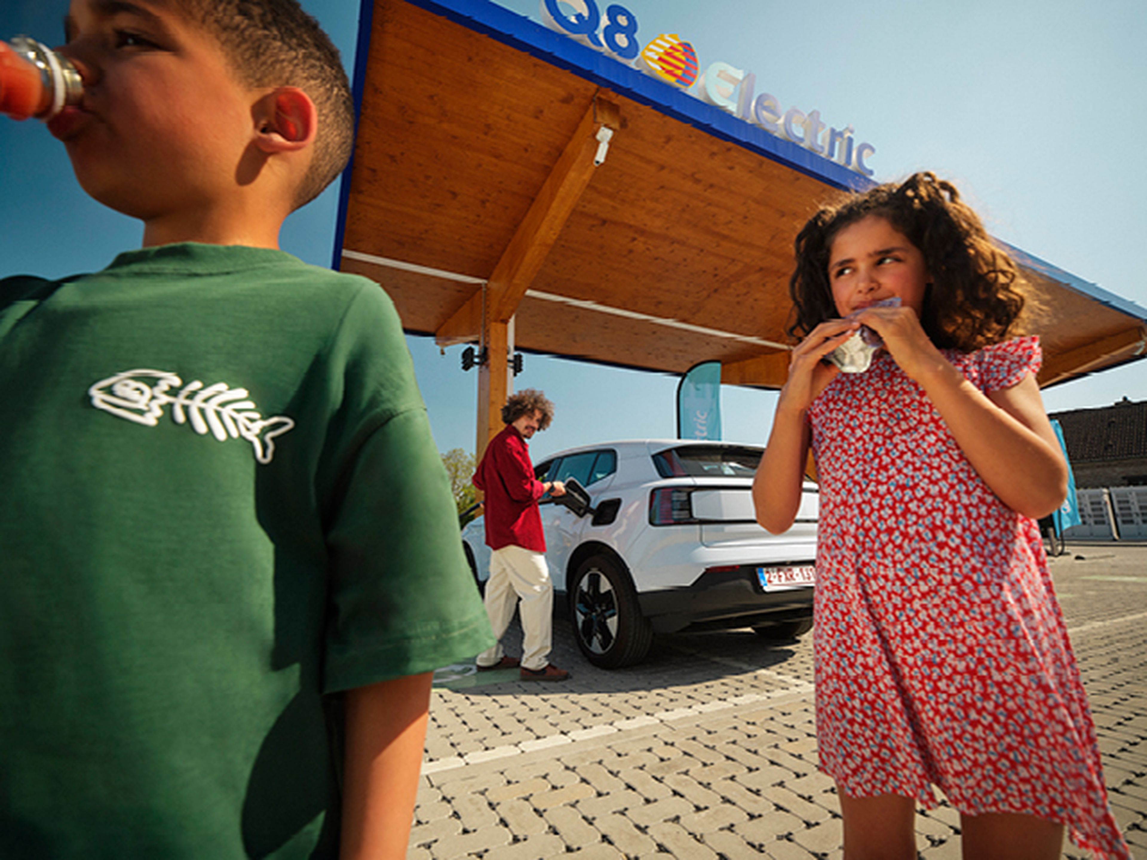 Kinderen drinken terwijl een man een elektrische auto laadt bij een Q8 Electric station.​