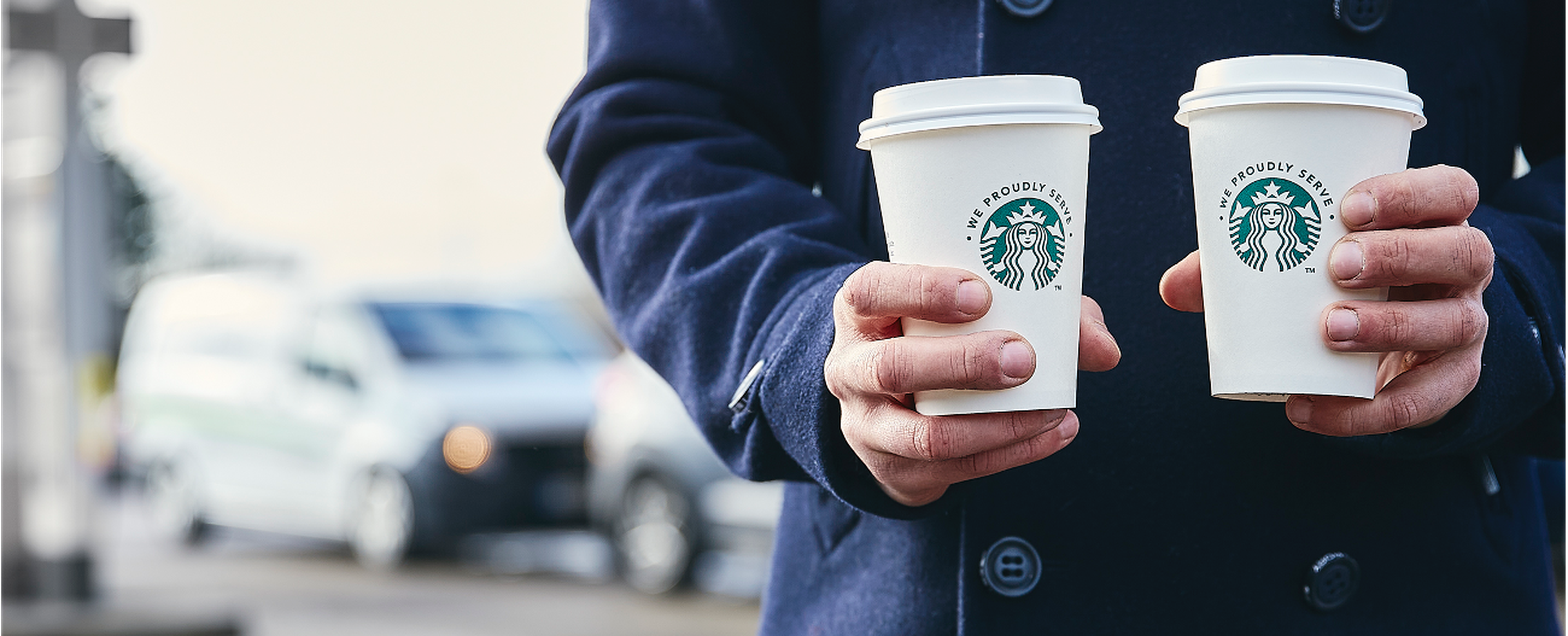 Personne en veste bleu foncé tenant deux tasses de café Starbucks dans un magasin Q8, avec des voitures floues en arrière-plan.