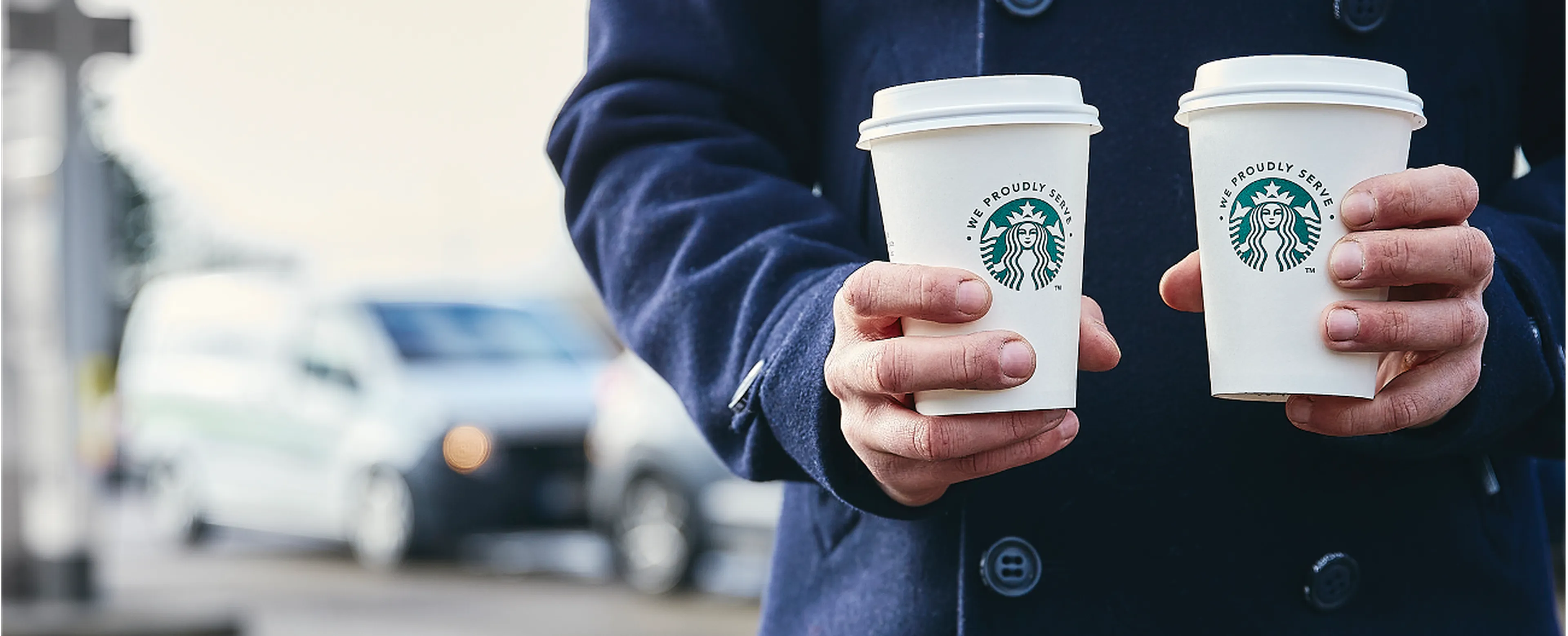 Persoon in een donkerblauwe jas twee Starbucks-koffiebekers vasthoudt bij een Q8 Shop, met wazige auto's op de achtergrond.