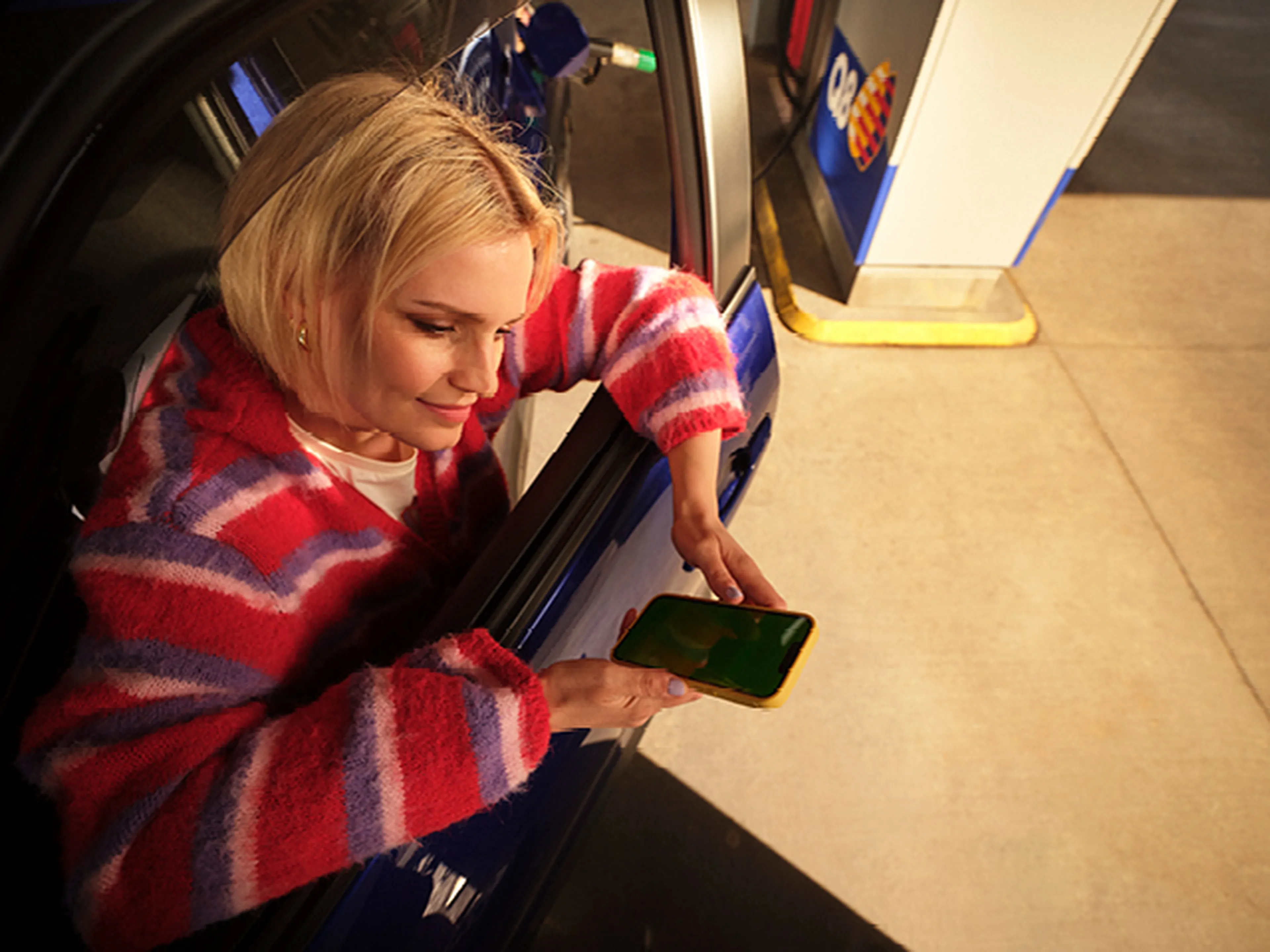 Een vrouw met blond haar en een kleurrijke gestreepte trui zit in haar auto bij een Q8 tankstation. Ze houdt haar smartphone uit het raam, klaar om iets te scannen of te betalen.