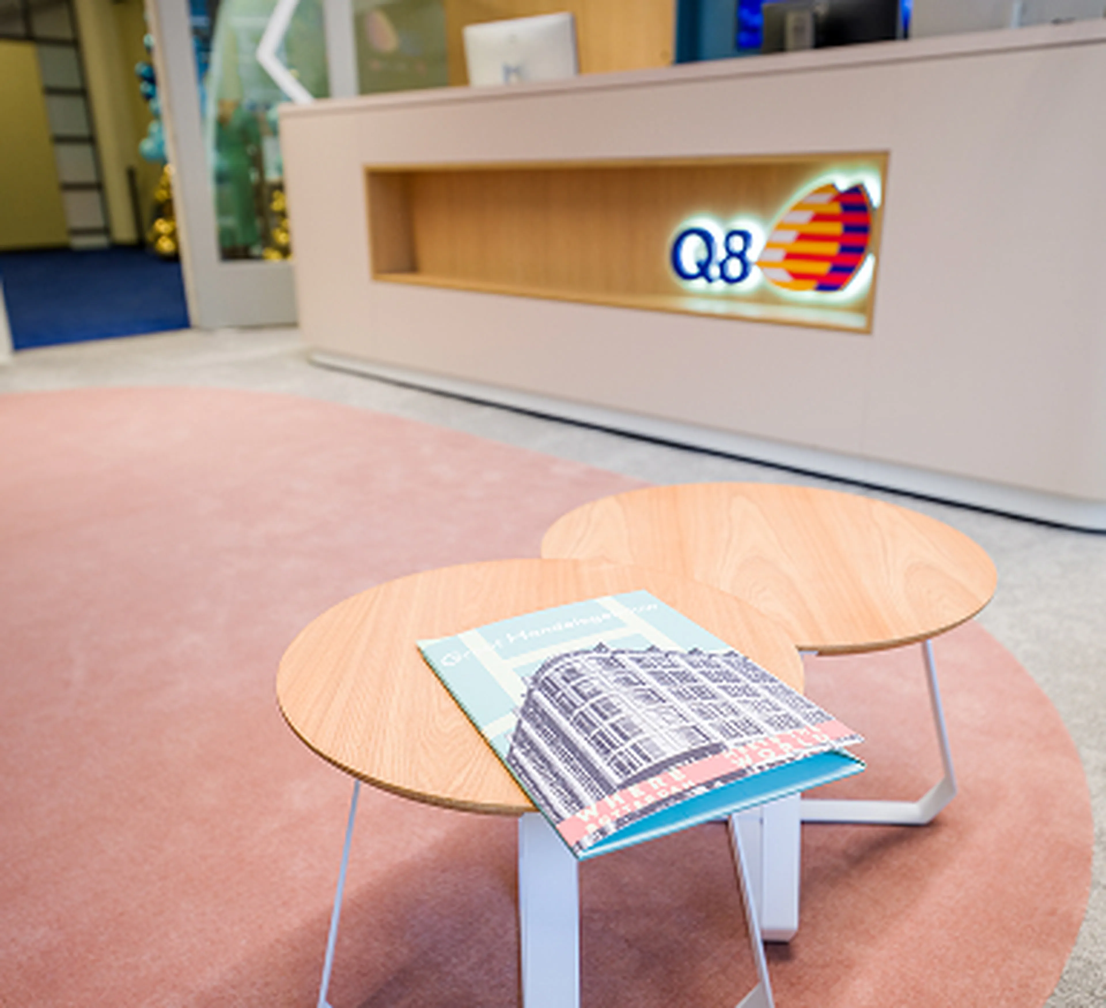 Reception desk of Q8 at the headquarters in Rotterdam, with the logo visible on the wall. In the foreground, two round wooden tables with a book on top stand on a soft pink carpet.