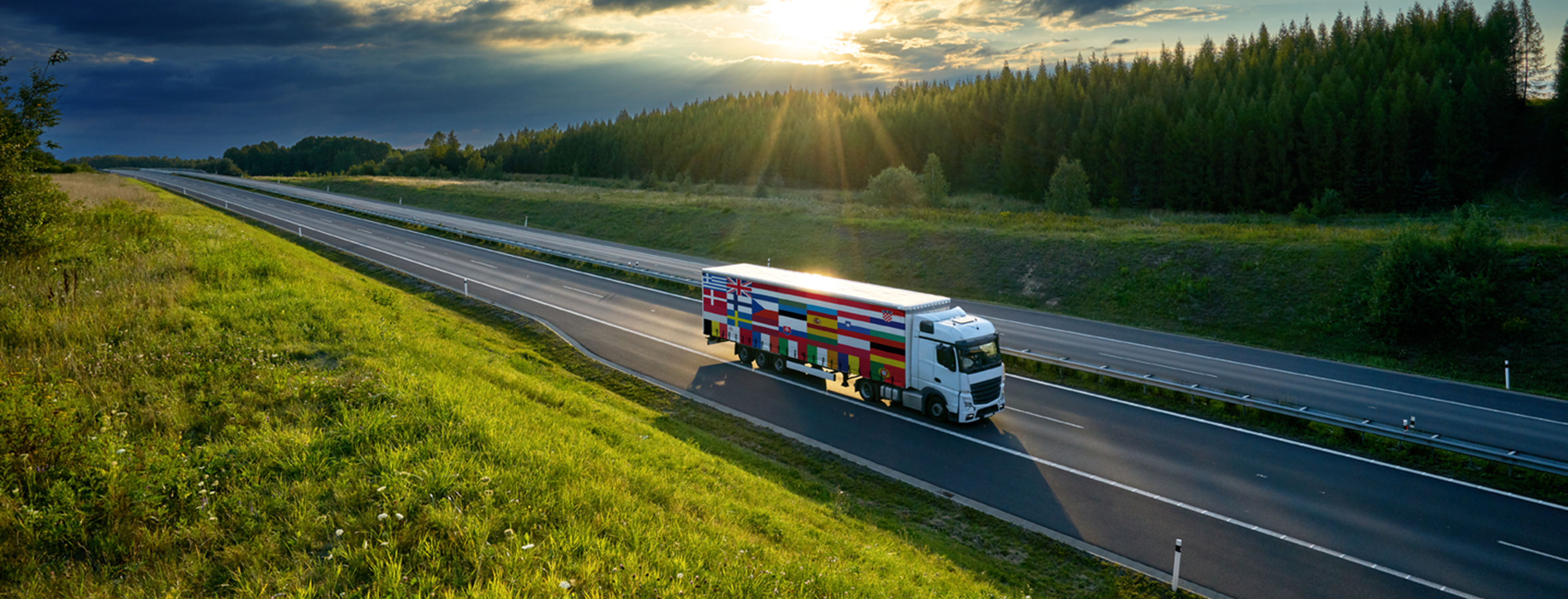 Truck with European flags on its side driving on an empty highway, surrounded by green grass and a forest edge, with sun rays breaking through the clouds.
