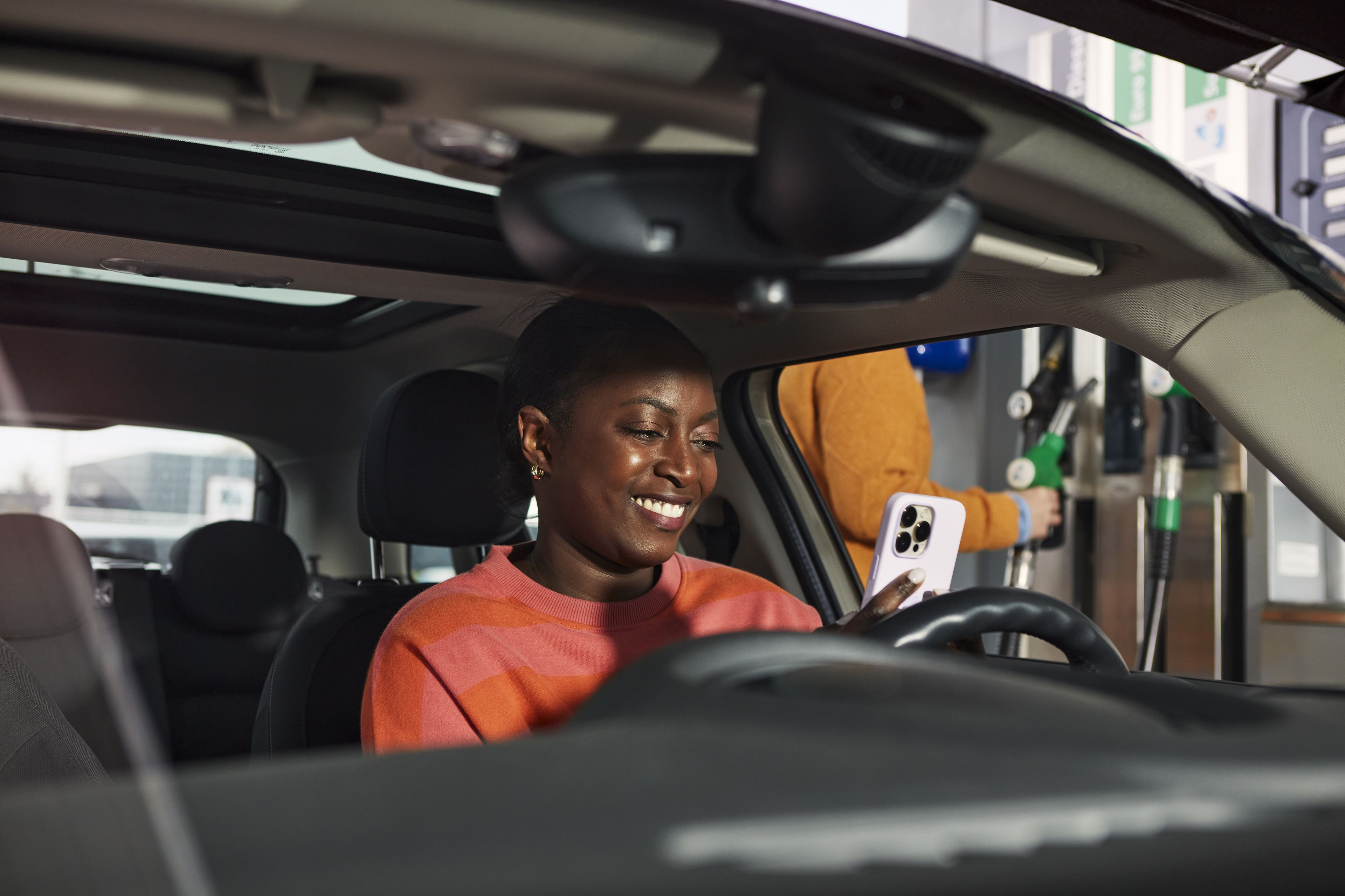 Woman in an orange sweater looks at her white smartphone while sitting in her car.