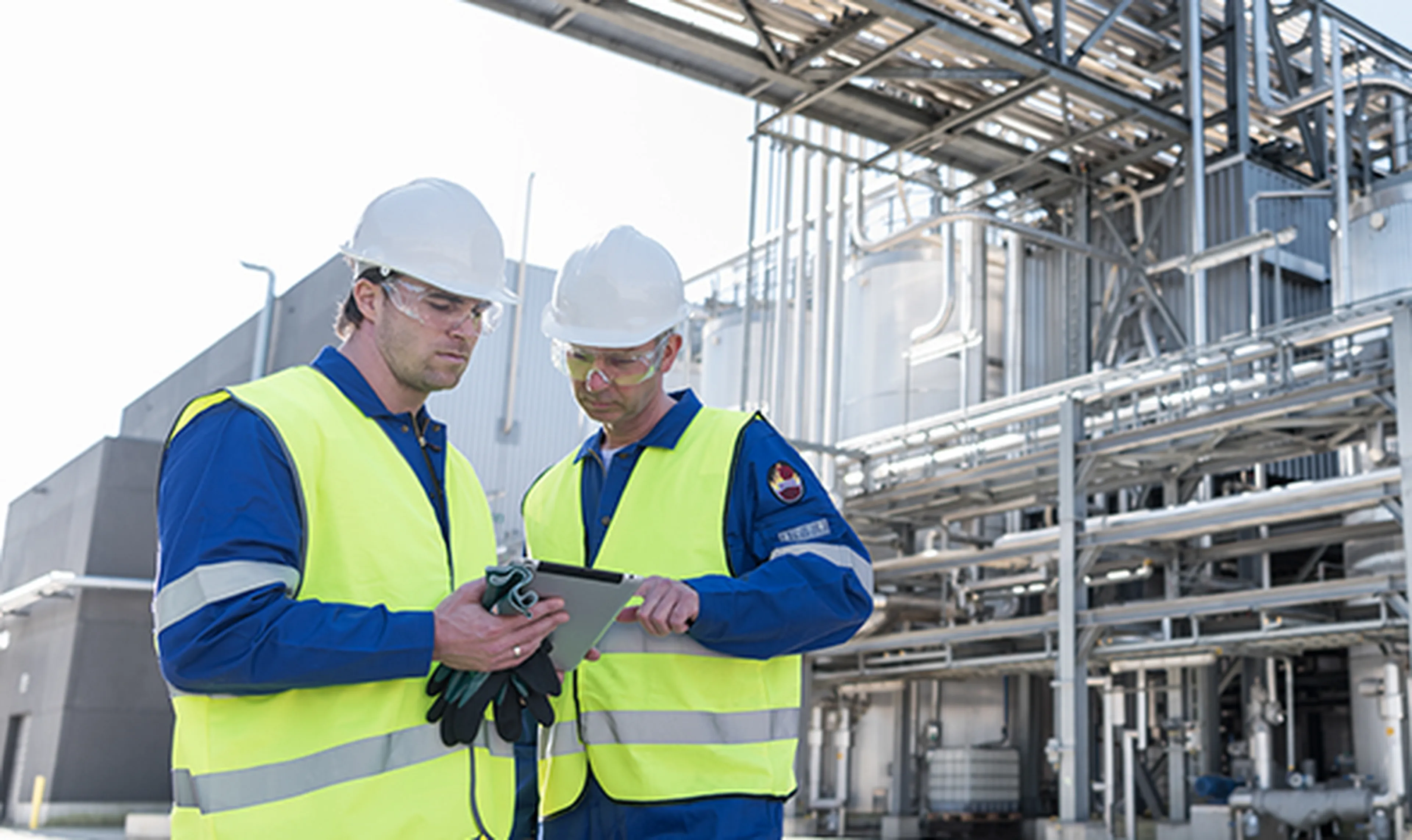 Two Q8Oils employees are wearing safety gear — blue overalls, yellow hi-vis vests, white helmets, and safety glasses — at an industrial site. In the background, metal pipes and installations from the blending plant are visible.