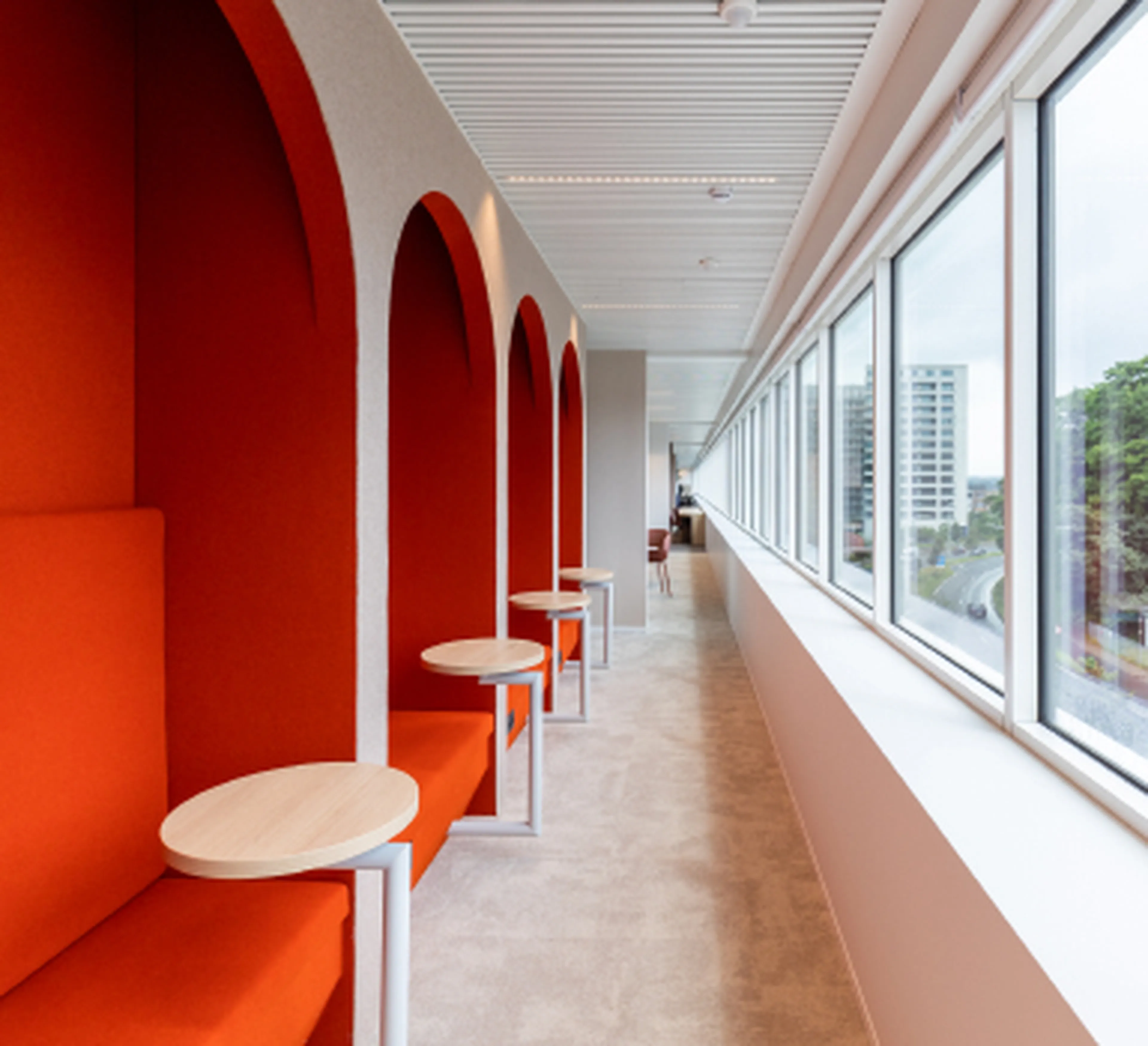 Long corridor with large windows at the Q8 headquarters in Antwerp. On the left are striking red alcoves with built-in benches and round tables, perfect for informal conversations or short breaks. On the right, a glass wall offers a view of the city.