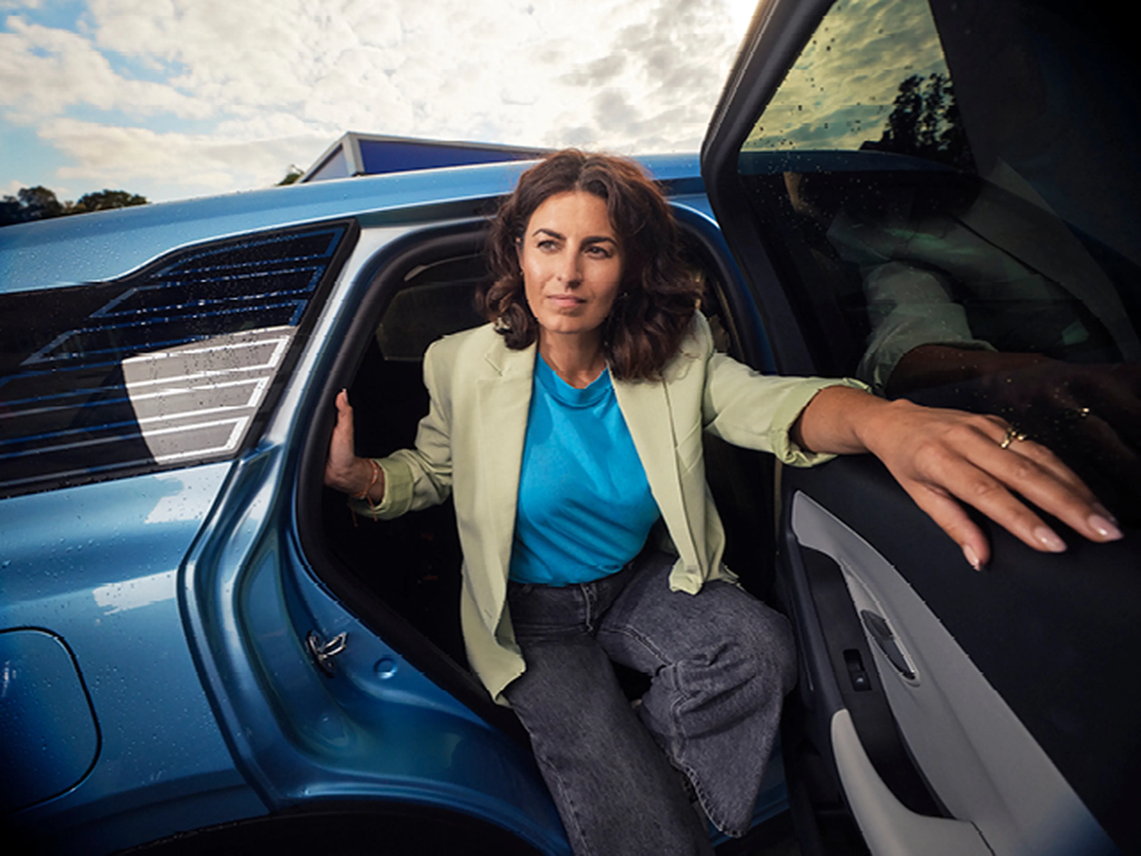 Een vrouw met donker krullend haar stapt uit een blauwe elektrische auto. Ze draagt een beige blazer, een felblauw T-shirt en een donkere jeans.